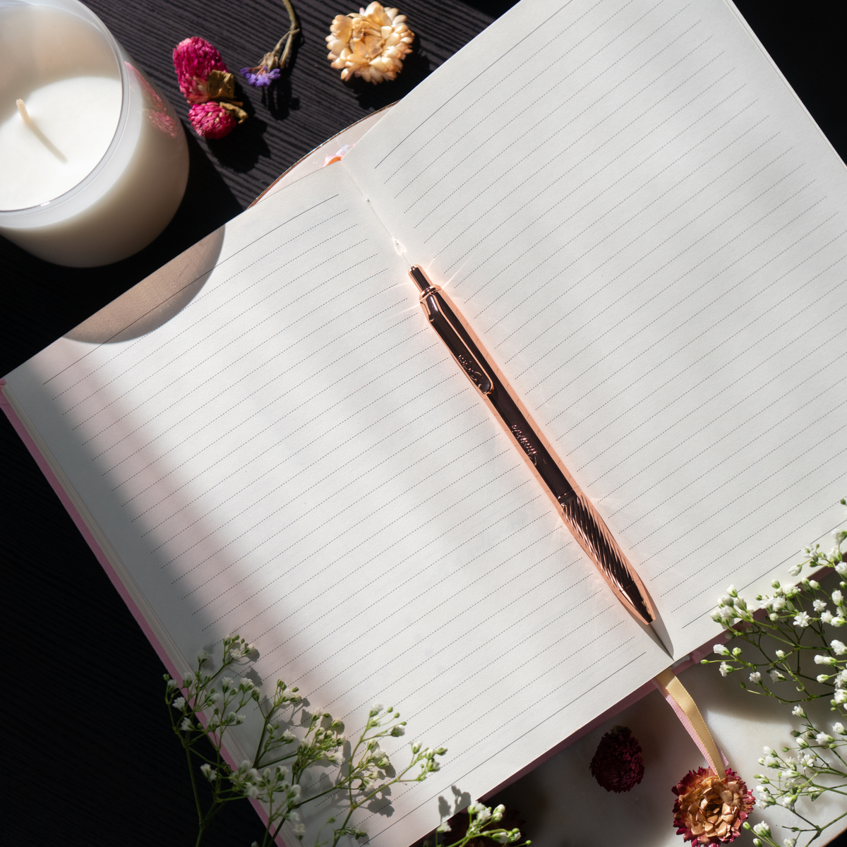 Rose gold pen on an open notebook with a candle and flowers in the background